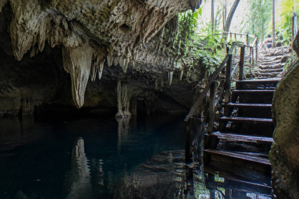 A wooden staircase leading down into a cave cenote with pale stalactites above dark still water