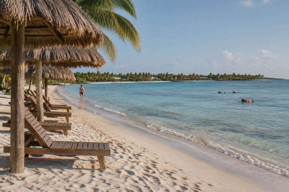 Rustic palapa shade structures on a sandy beach with calm turquoise water and a distant headland under a clear sky