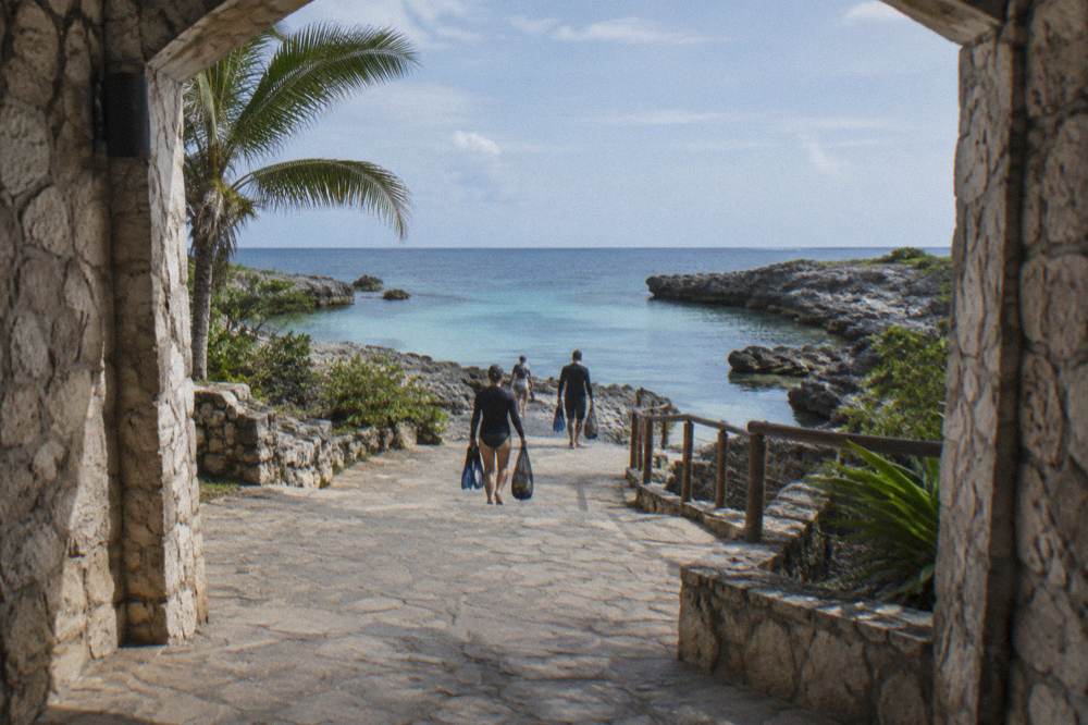 Tour group with snorkel gear walking from a beach entrance toward calm morning water