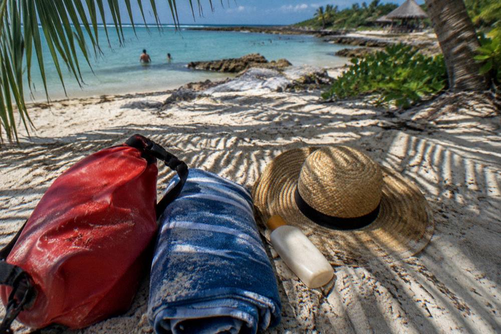 A dry bag, towel, sunscreen bottle and hat arranged on sand under palm shade