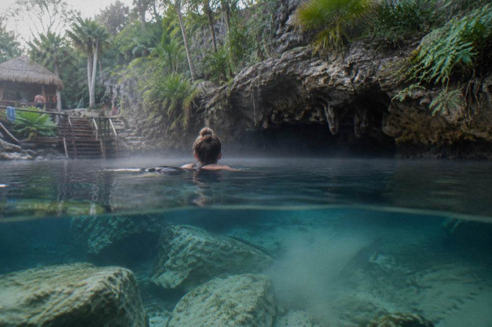 A swimmer at the surface of a cenote near a cave entrance with underwater rock visible