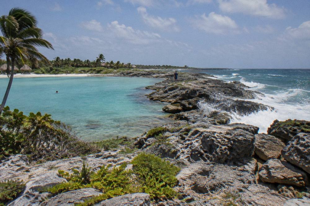 A rocky point separating two bays, with calm turquoise shallows on one side and gentle waves on the other.