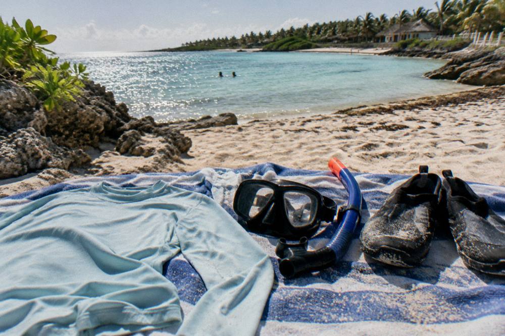 A sun shirt, mask, snorkel and water shoes laid out on a beach towel beside a crescent bay shoreline.
