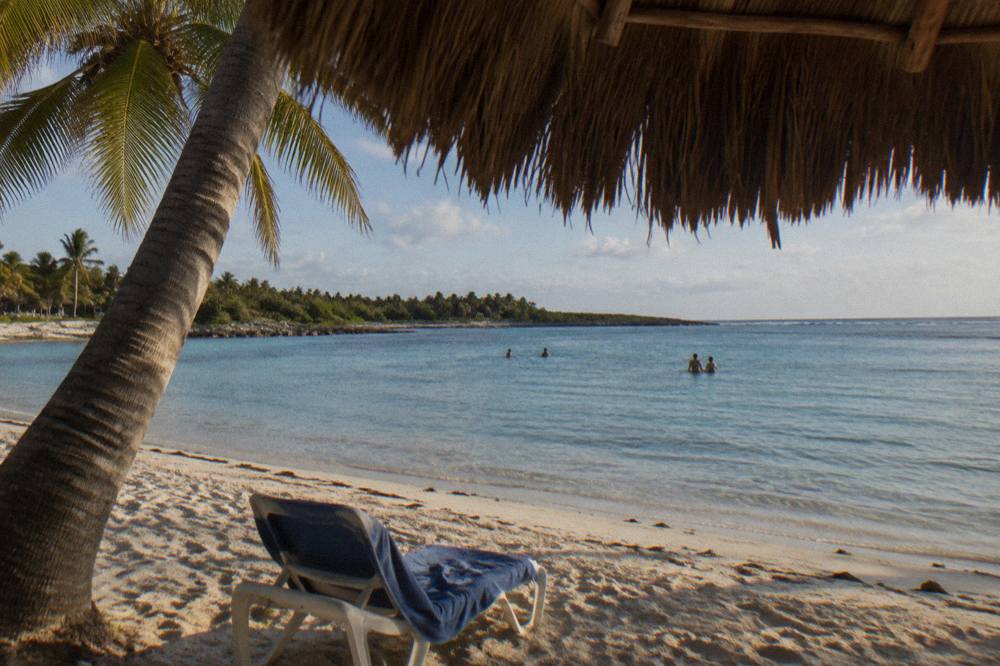 A palm-fringed white sand beach with calm shallow water and a wooden palapa roof