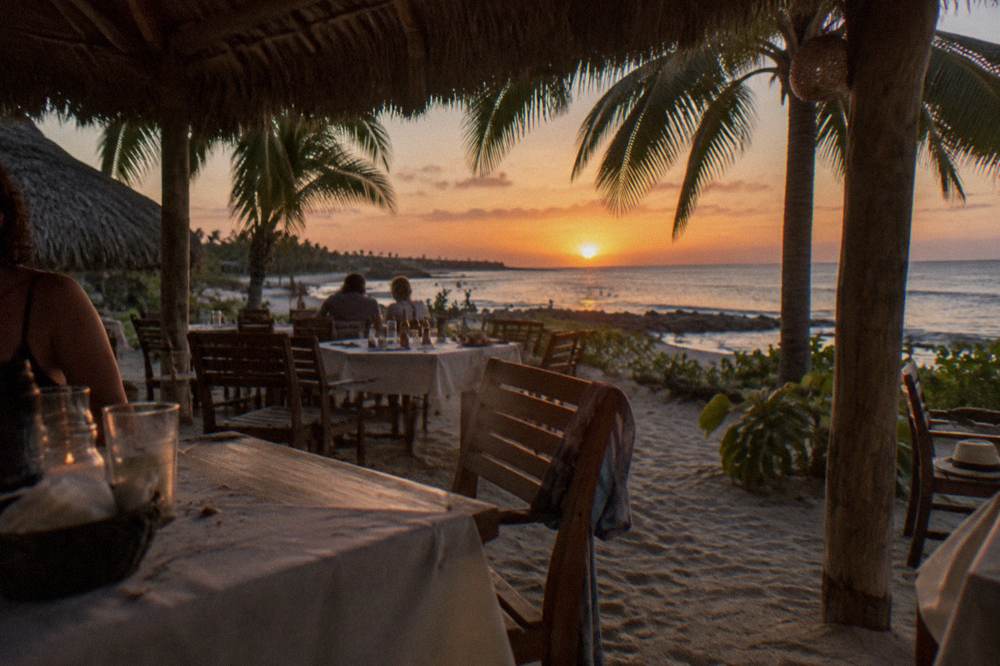 An open-air beachfront restaurant terrace with tables set on sand under palm thatch
