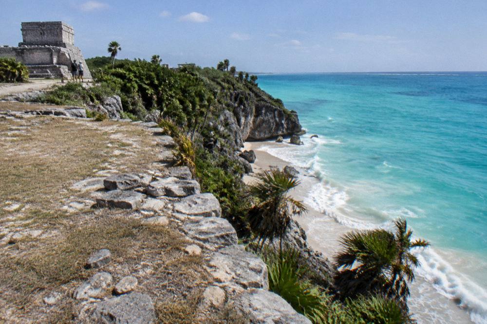 A cliffside stone ruin overlooking a turquoise sea with a sandy cove below