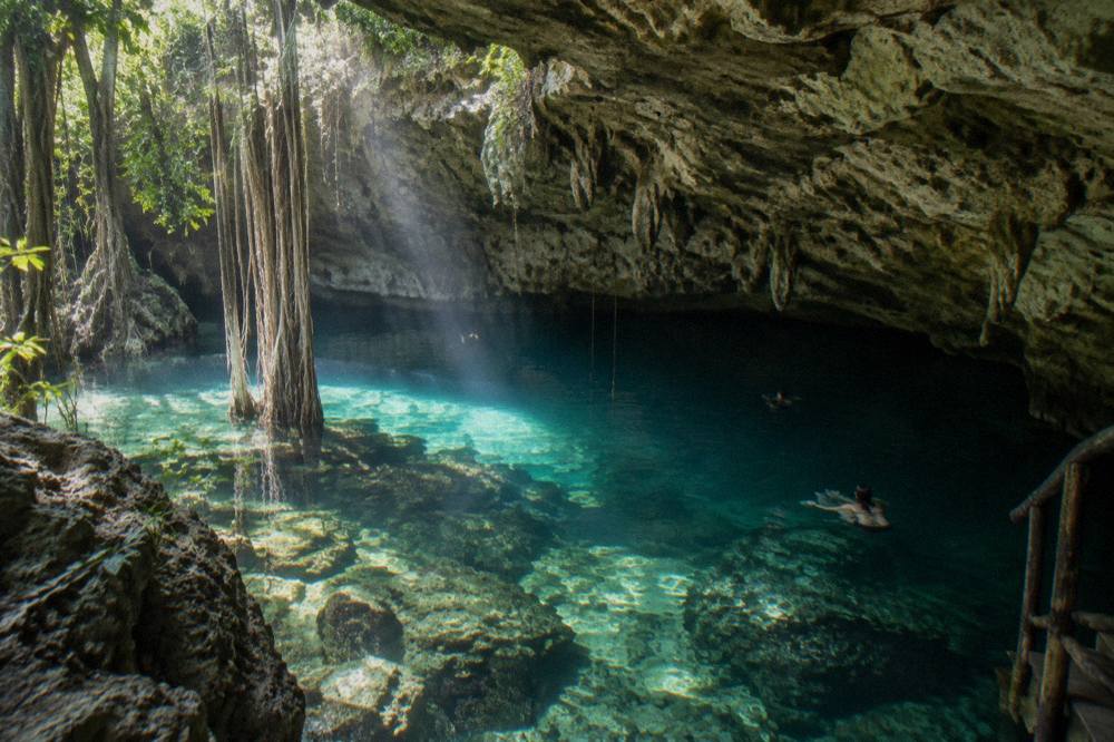 A limestone cenote with clear water, overhanging rock and sunlight entering through an opening