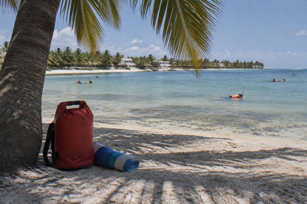 A dry bag and towel resting on warm sand under a palm tree near clear shallow water