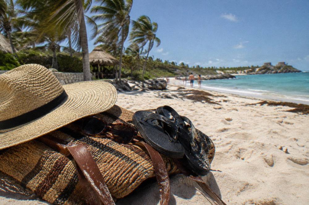 A wide-brim hat, sunglasses and sandals arranged on a beach bag under palm shade