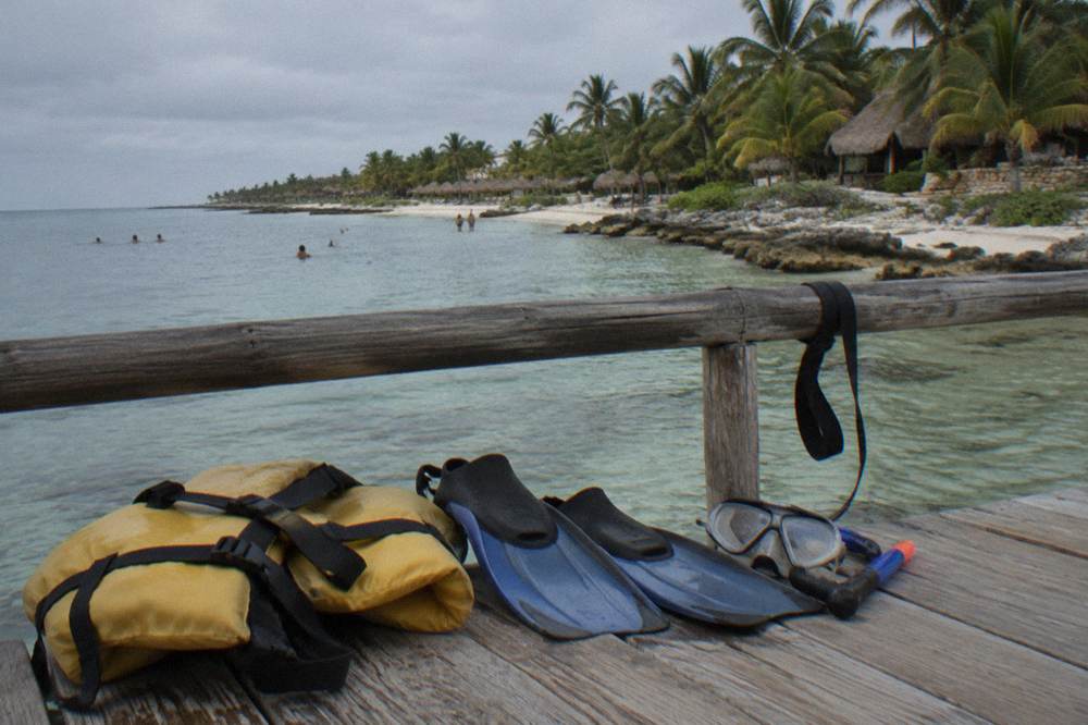 A life vest, short snorkel fins and a mask laid out on a wooden bench beside a calm bay