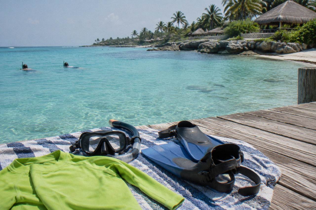 Snorkel mask, fins and a rash guard laid out on a beach towel beside calm turquoise water