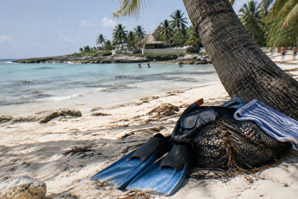 Snorkel fins, a mask and a folded towel laid out on warm sand beside a mesh bag